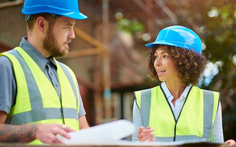 Two people in hi-viz tabbards and blue hard hats holding clipboards and discussing matters on a building site.