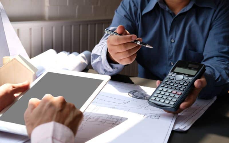 Two people working out the cost of building a home. The plans are set out on a table and the two men are working with a calculator and a tablet device to estimate the costs.