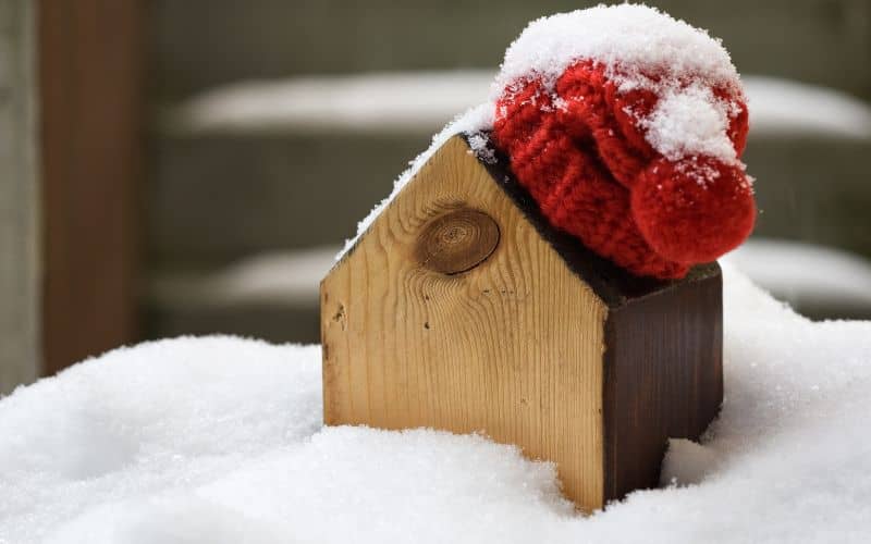 A wooden model house sitting in snow with a red woolly hat on its roof.