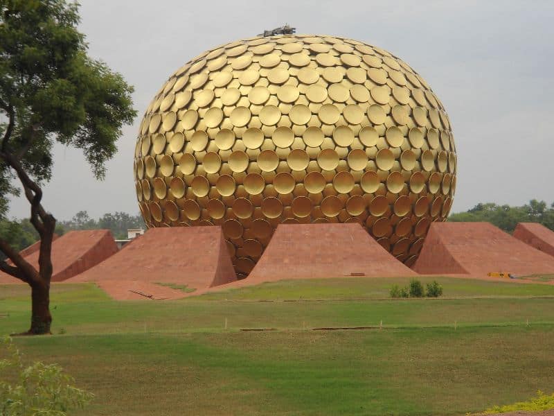 A large ovoid-shaped building called Matrimandir. It has golden discs all over its outer surface and earthen construction surrounding it. The structure is set in parkland.