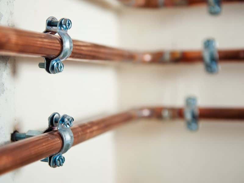 Copper pipes running along a wall in parallel. The pipes are held in position on the wall by steel clips.