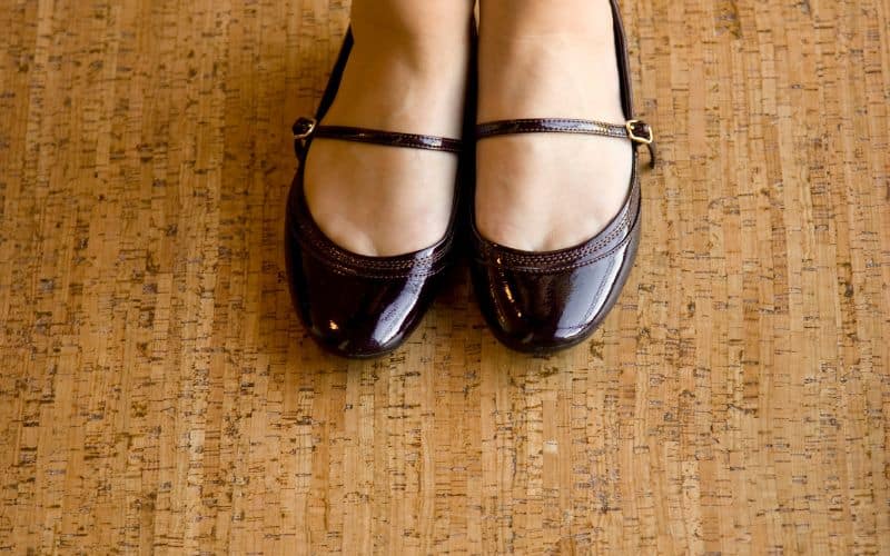 A woman's feet, wearing smart shoes with no socks, standing on cork flooring.