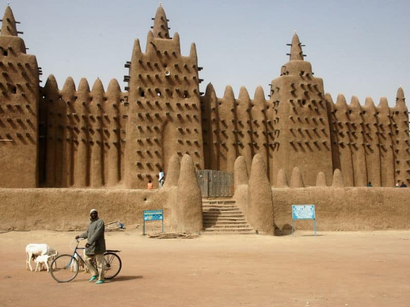 An image of the Great Mosque of Djenné. It is a large structure made from rammed earth with pieces of wood that stick out of it at regular intervals that serve as built in scaffolding. There is a set of steps at the front of the mosque that lead up to the main wooden door.