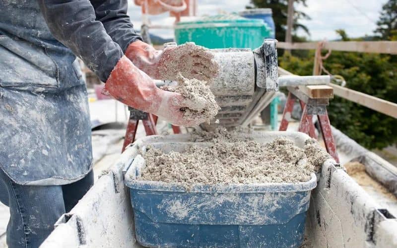 A worker mixing hempcrete by hand on site. The worker is wearing red protective gloves and a blue apron.