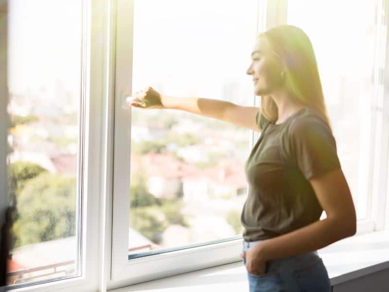 A woman with long hair opening a window in a brightly lit home.