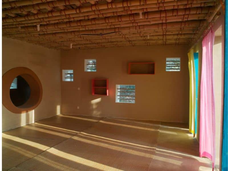 A room made from earth. The rammed earth walls and floor are brown in color. Windows are set into the wall and brightly colored curtains hang along the right hand wall.