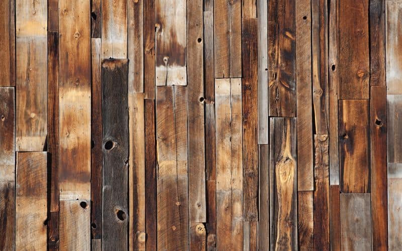 Lots of planks of reclaimed wood leaning against a wall. The wood is of various colors, ranging from sanded, natural wood do deeply-stained, dark brown pieces.