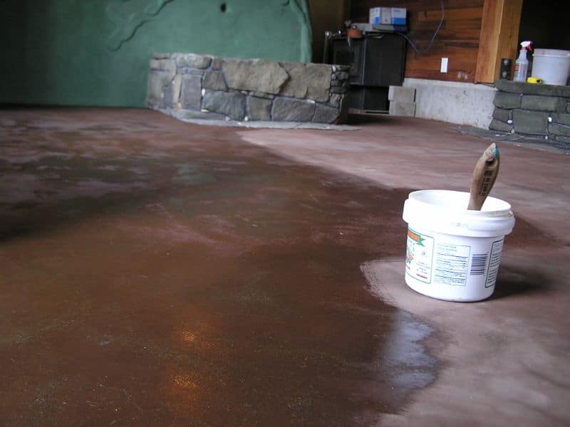 An earthen floor half painted with linseed oil. There is a pot of linseed oil with a paintbrush sticking out of it next to the painted area. Stone features and a green painted wall are visible in the background.