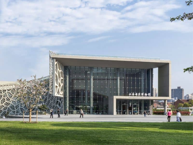 An image of the Shanghai Natural History Museum in Shanghai, China. It is a cuboid shape, which is fully glazed and has a large paved area in front with a green lawn beyond. There is a lattice structure to the left, which is joined to the main building.