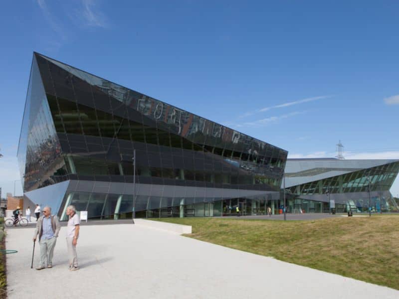 An image of The Crystal, London, UK. The building is mainly tinted glass. Its shape resembles the prow of a boat and there is a large paved area and a grass lawn out front.