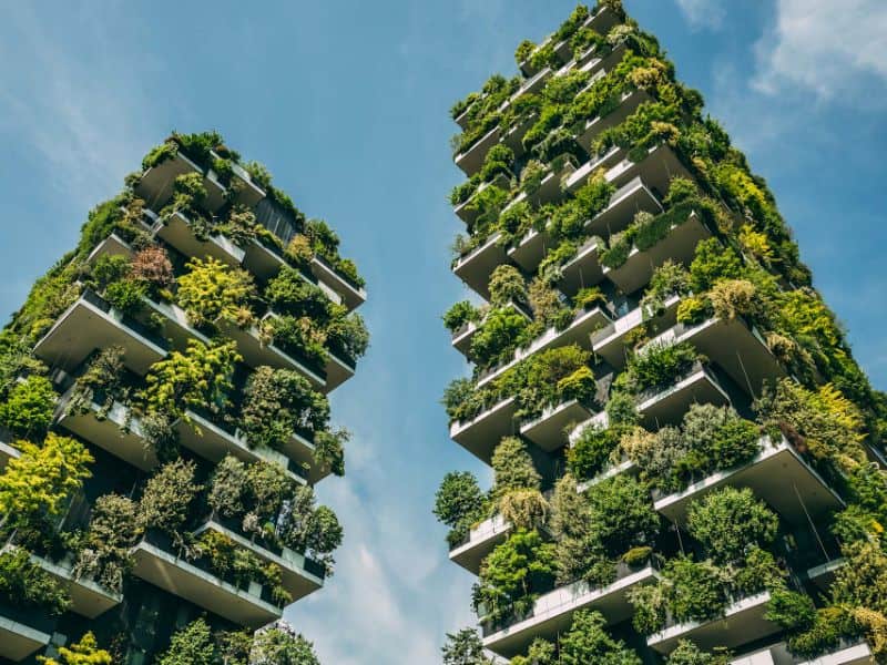 An image of the Vertical Forest, Milan, Italy. This is a pair of towers largely covered in shrubs and greenery. They are set against a blue sky background.