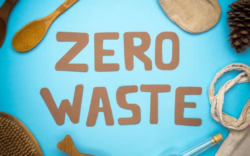 The words "zero waste" written on a bright blue background. There are a number of implements and containers made from wood and other natural materials surrounding the text.