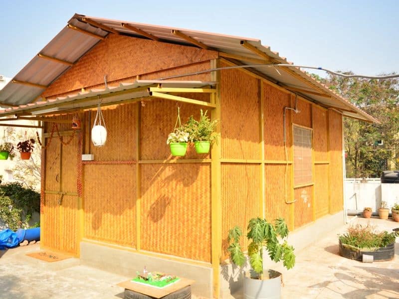 A bamboo hut with a corrugated metal roof and porch. The house is on a concrete slab foundation and has potted plants handing from its eaves and in pots around it.