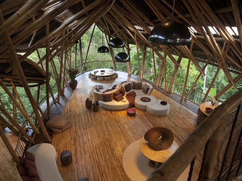 A stylish bamboo home viewed from the inside. The roof members are visible and the sides of the home are open, creating a spacious, airy living area. There is bamboo flooring and an S-shaped sofa in the middle of the room with a circular table and stools at the far end.