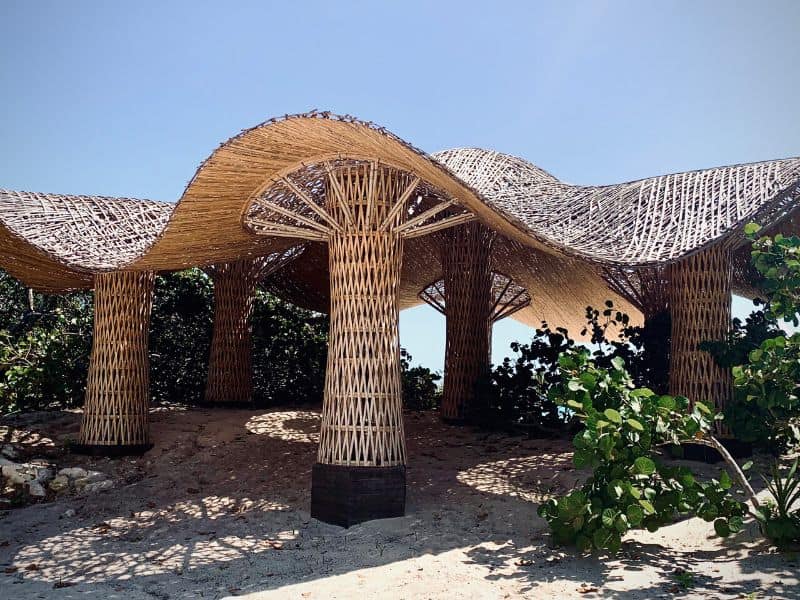 A woven bamboo structure comprising bamboo pillars and a curved roof set on sandy ground. The sky above is pure blue and there is green vegetation in the background.