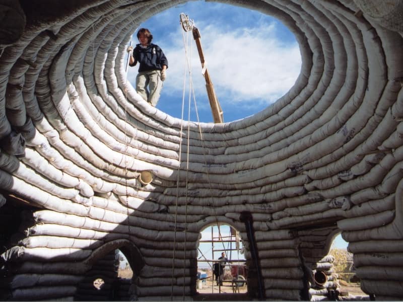 The CalEarth EcoDome, which is a house made from earthbags in a dome shape. The dome is unfinished and viewed from inside looking up. A worker can be seen working at the top of the dome against the blue sky with whispy white clouds. There are openings for windows and doors, through which