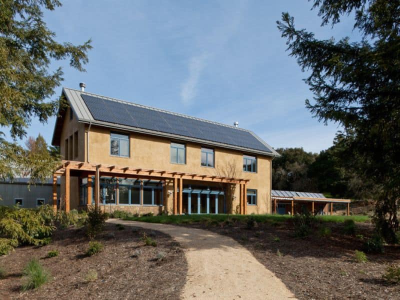 A two-story straw bale house with a pitched roof with solar panels mounted on it. There is a large pergola out front and many windows in the elevation facing the camera. There is a dirt path curving gently up to the front door.