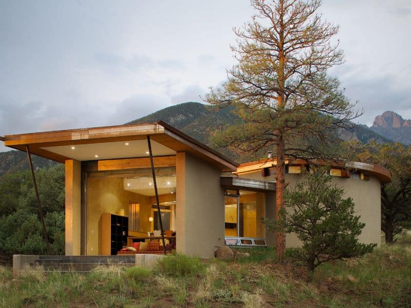 A photograph of a straw bale house of modern design. It is single story and has flat roofs. There are three distinct sections to the house - one square, one circular, and a linking corridor. It has a wooden frame and large glass windows. There are mountains in the background.