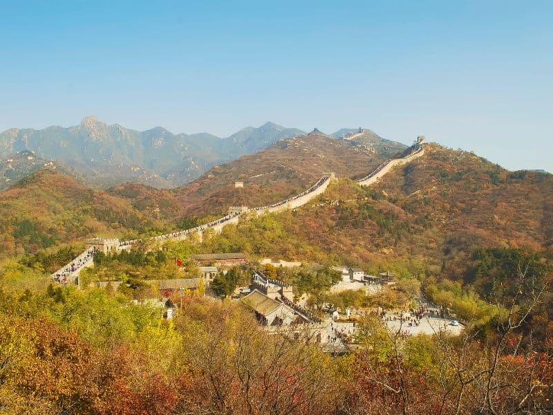 A picture of a section of the Great Wall of China with mountains in the background, trees in the foreground and a small settlement in the center of the frame.