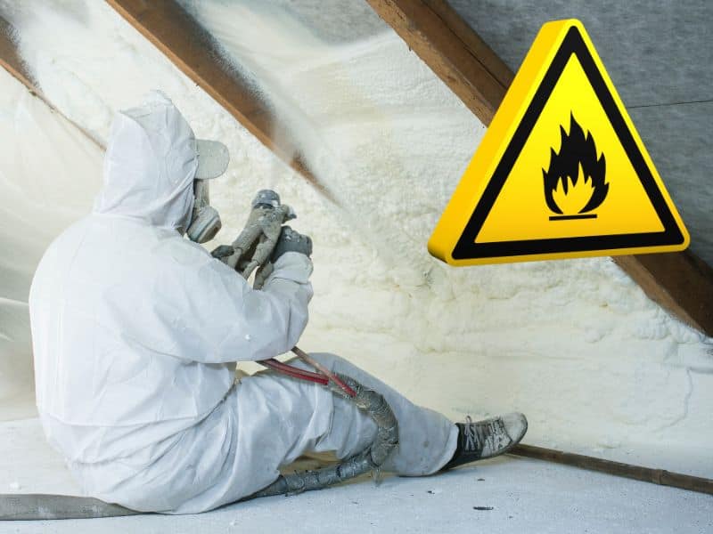 A picture of a worker installing spray foam using a spray gun between rafters in a roofspace. There is a black-on-yellow flammable warning sign overlain.