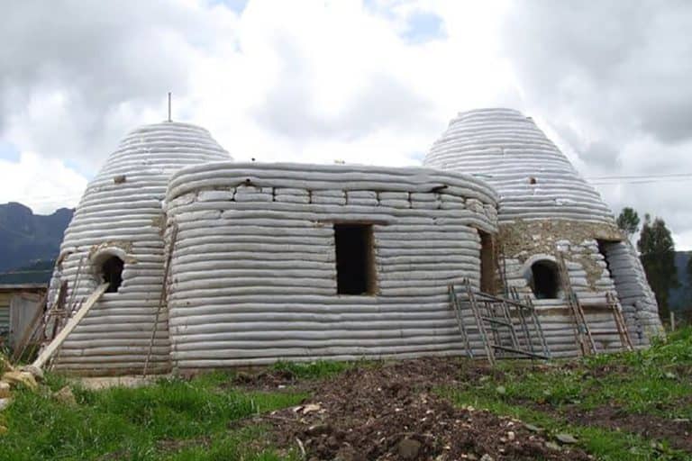 An earthbag home comprising two dome structures either side of a cuboid shape with rounded corners. The domes have circular holes for the windows and the cuboid shaped section has rectangular holes. The house is still under construction and the layers of earthbag can be clearly seen.