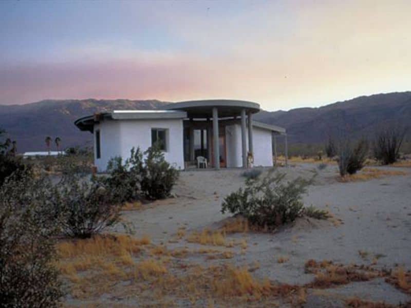 A single story straw bale house with white walls and a small porch area at the front. The house grounds are sandy with sparse vegetation and mountains in the background.