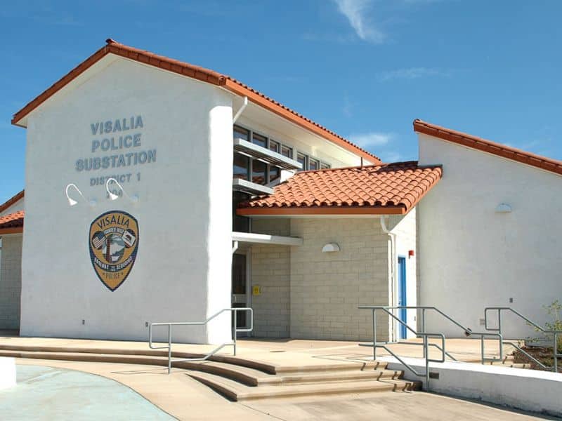 A photograph of the Visalia Police Substation in Visalia, California. The building is multi-story and has orange clay roof tiles. The walls are painted white and there is police livery on the gable end. Wide steps lead up to the front door with railings either side.