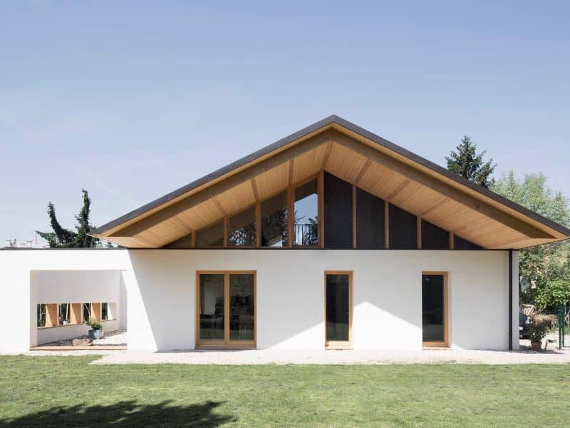 A two-story straw bale house with a pitched roof and tall windows. The window frames are light-colored wood, which matches the underside of the roof. The walls are white and there is a green lawn out front.