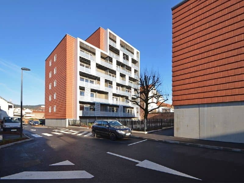 A seven-story apartment block with balconies for each apartment and orange cladding on the end of the building.