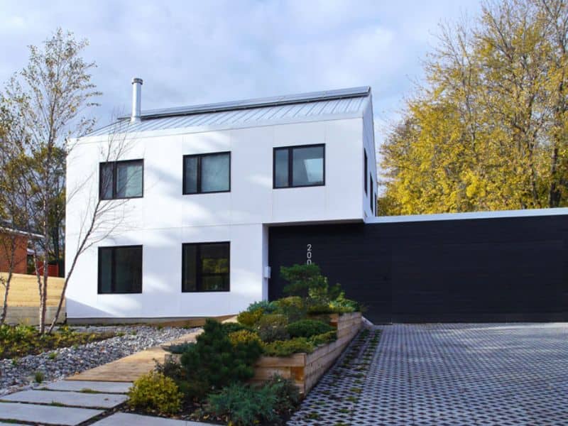 A two-story straw bale house with a corrugated metal roof and black window frames. There is a black-clad annex on the ground floor and permeable paving in the driveway.