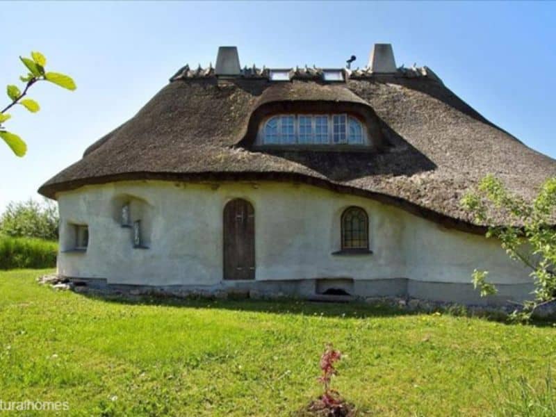 A characterful straw bale home with cream-colored walls. The building is roughly round in plan and has a brown thatched roof. The front door is narrow and wooden with a rounded top. There are many irregular-shaped windows with small panes of glass.