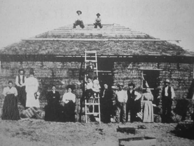 An old, grainy, black and white photograph of the load-bearing style of straw bale construction that was popular in Nebraska in the early 1900s. The house is single story and has the roof in place but not yet finished. There are two men sitting on the ridge of the roof with a ladder leaning against the front wall. There are 13 people lines up on the ground in front of the house posing for the camera.