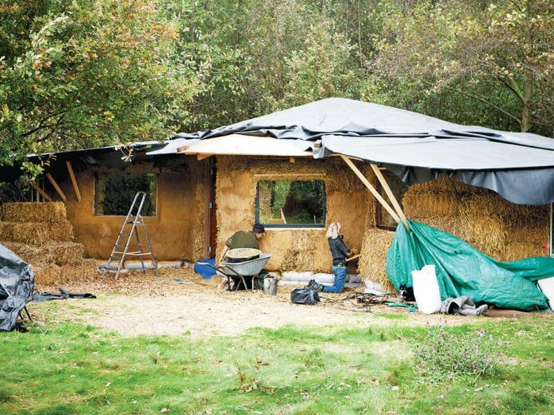 A single-storey straw bale house under construction. The roof is partly completed and the walls are being plastered on the outside. There are two people working on the house and there is a wheelbarrow and set of step ladders in view.