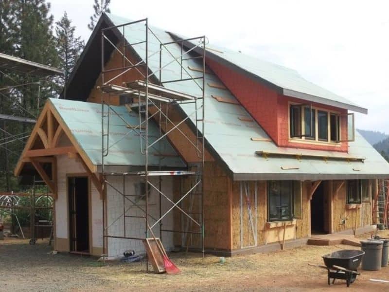 A straw bale house being built by Many Hands Builders. It's a two-story home with a large dormer window and steeply-pitched roof. The breather membrane is visible on the roof, and the straw bales can be seen in the walls as the home is only partially completed. There is a single-story porch on the gable end and some scaffolding leading up to the roof.