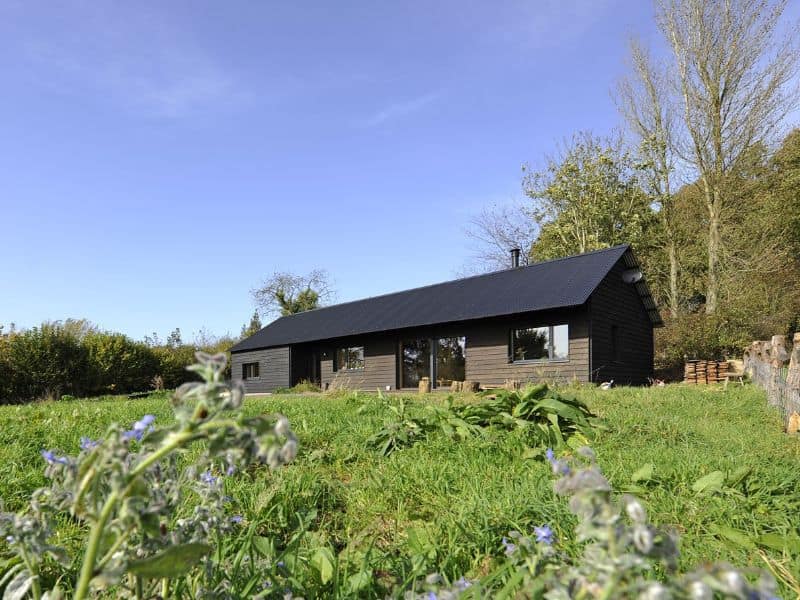 A single story straw bale house with dark wood cladding on the walls and a black corrugated metal roof. The window frames are dark gray and there is a pair of sliding glass doors in the middle of the front elevation. There is a wood-burning stove flue coming out of the top of the roof. The house is set in a field of grass with green hedgerows around the edge.