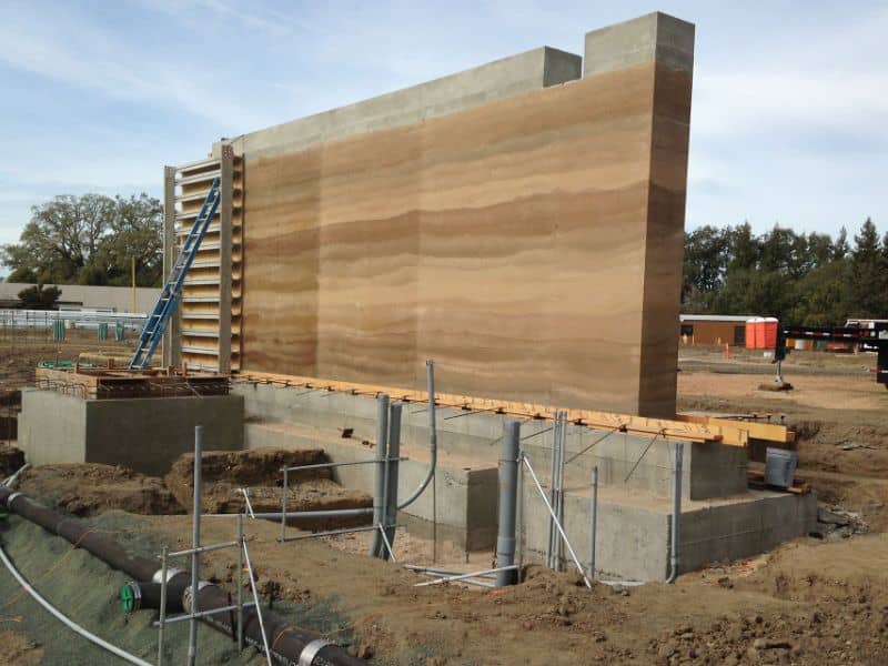 A rammed earth wall under construction. The concrete block footings are clearly visible as is a lot of ducting and pipework in the ground next to the wall.