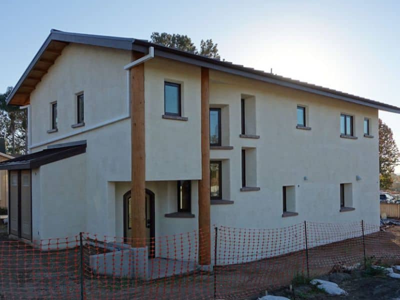 A large two-story straw bale house with small windows place in a somewhat irregular pattern along its walls. There are two large wooden pillars either side of the porch that support the upper floor. The window frames are black and the walls are cream colored..