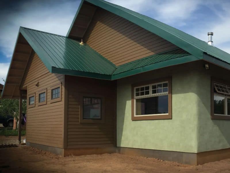 A sage and green straw bale house with wood panelling on the gable end. The ground surrounding the house is bare earth, indicating that the house has recently been built.