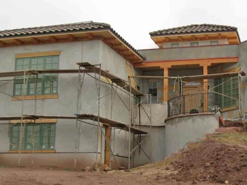 A two-story straw bale home that has been freshly plastered. It has a tiled roof, scaffolding around the outside and bare earth grounds.