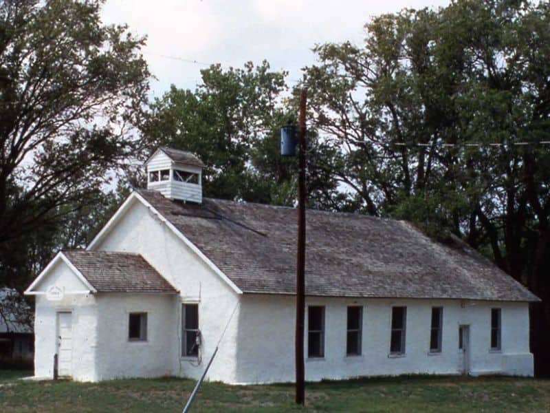 A photograph of the Pilgrim Holiness Church in Arthur, Nebraska. The church is a single story building with white walls and a slate roof. There is a bell tower at the top and a small porch on the end.