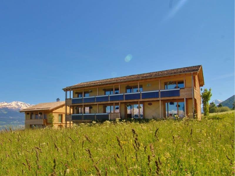 A straw bale hotel set in a beautiful meadow. The walls are light yellow and there is a long balcony that runs all the way along the upper floor. There are snow-capped mountains in the background.