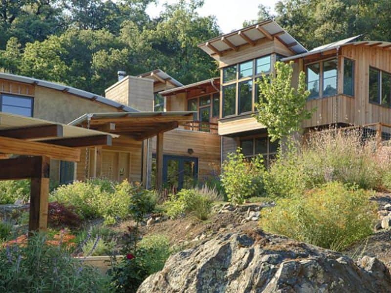 A straw bale house with wood cladding over parts of it. There are large windows and the house is arranged in a series of interconnected rooms over several levels, set on rocky ground.