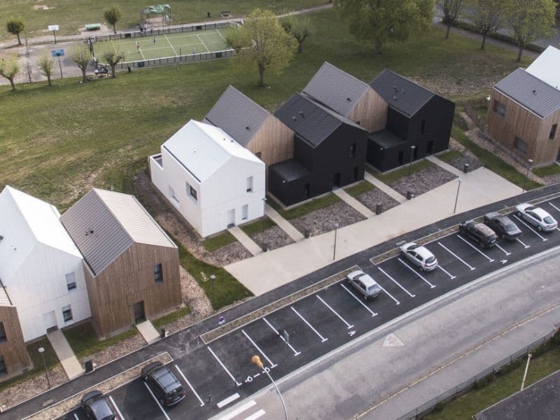 A small housing development made from wood and straw bales. The houses have staggered frontages, with every other house set back from the rest, which creates a small private area that is not overlooked. There is parking out front and footpaths linking all the houses together. The houses are different colors, with some being predominantly black, others natural wood color and others white. There is a small sports facility behind the houses in parkland.