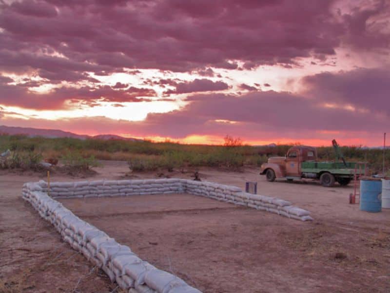 The first few layers of earthbags forming the walls of an earthbag house in the desert. There is a truck parked a few feet away and vegetation beyond the foreground of the image.