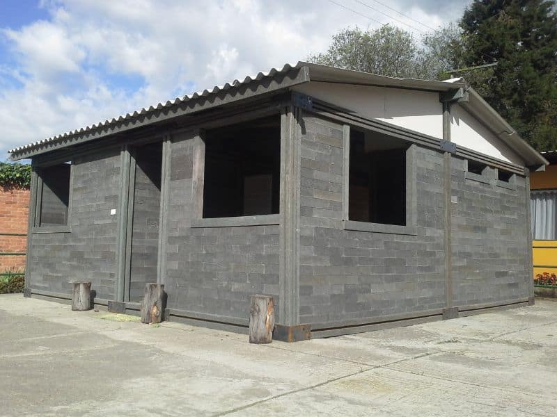 A house built from recycled plastic bricks by Conceptos Plásticos in Colombia. The walls are dark gray in color and the house has a corrugated metal roof. The windows have not yet been fitted but the holes in the walls are rectangular. The house sits on a concrete slab that extends away from the walls to form a large paved area.