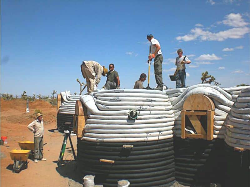 An earthbag house part way through construction with many workers building it. The bottom halves of the walls are black and the top halves are white.