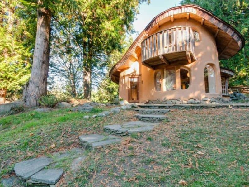 A beautiful cob home set in woodland. The walls are curved and smooth. The roof is curved and there is a first floor balcony.