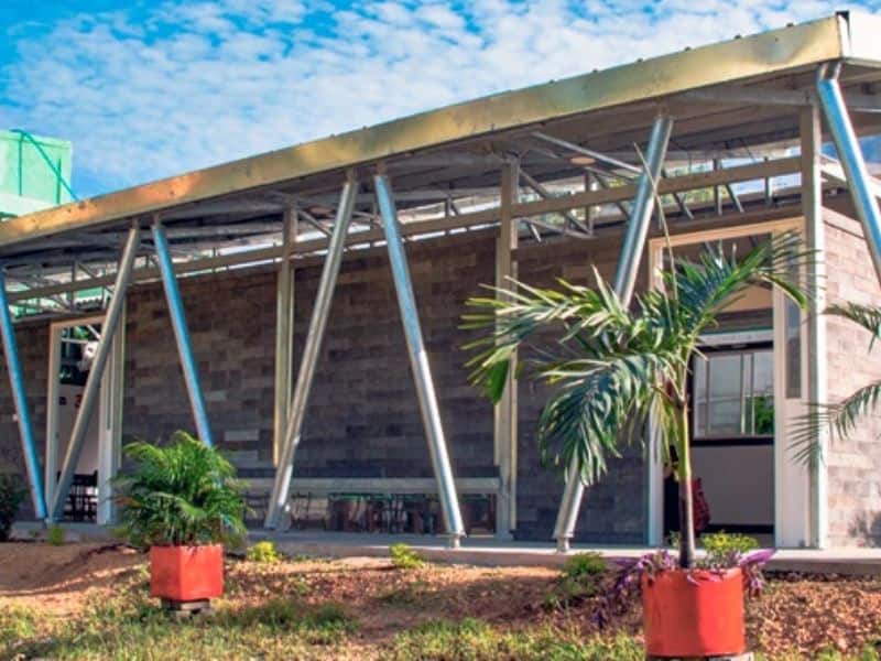 A building made from the recycled plastic blocks produced by Conceptos Plasticos. The blocks are dark gray and the roof is supported by criss-cross metal struts. There are palm trees in orange plant pots either side of the image.