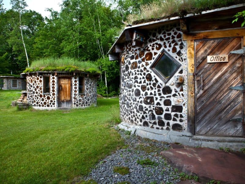 Cordwood buildings set in woodland. The roofs are covered in grass and the buildings themselves are circular with wooden doors and small windows.