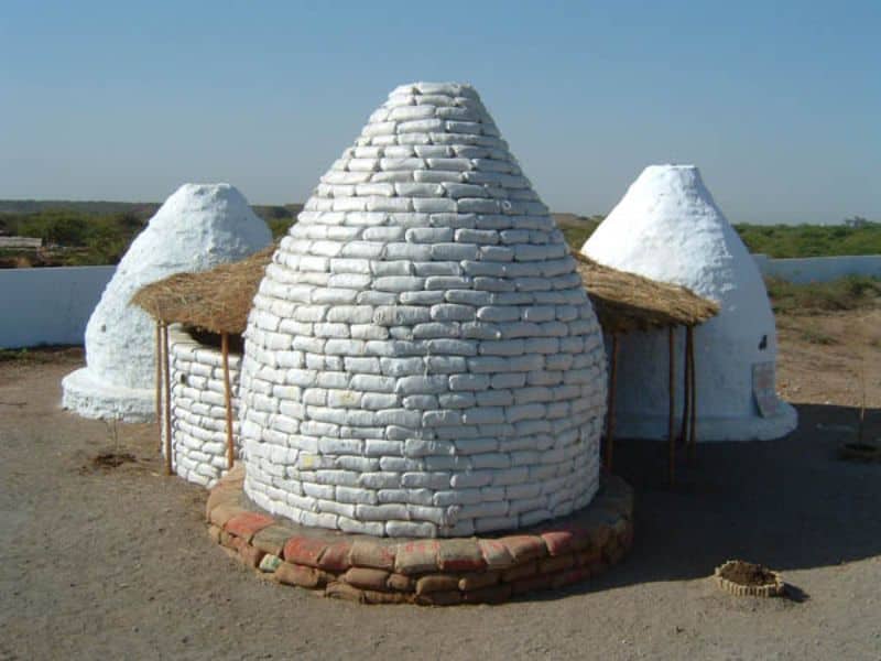 An earthbag building constructed from white earthbags. The building comprises three domes and a central, circular room with a thatched roof.